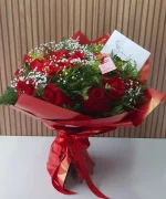 Buquê de rosas vermelhas com gipsofila branca e folhagens verdes, embalado em papel vermelho com laço decorativo