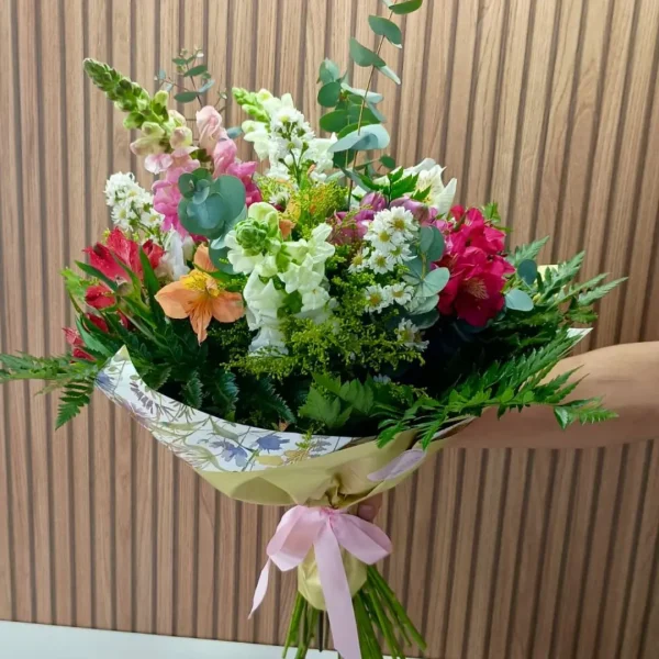 Buquê Bela de flores mistas coloridas com folhagens verdes, embalado em papel decorativo e laço rosa.